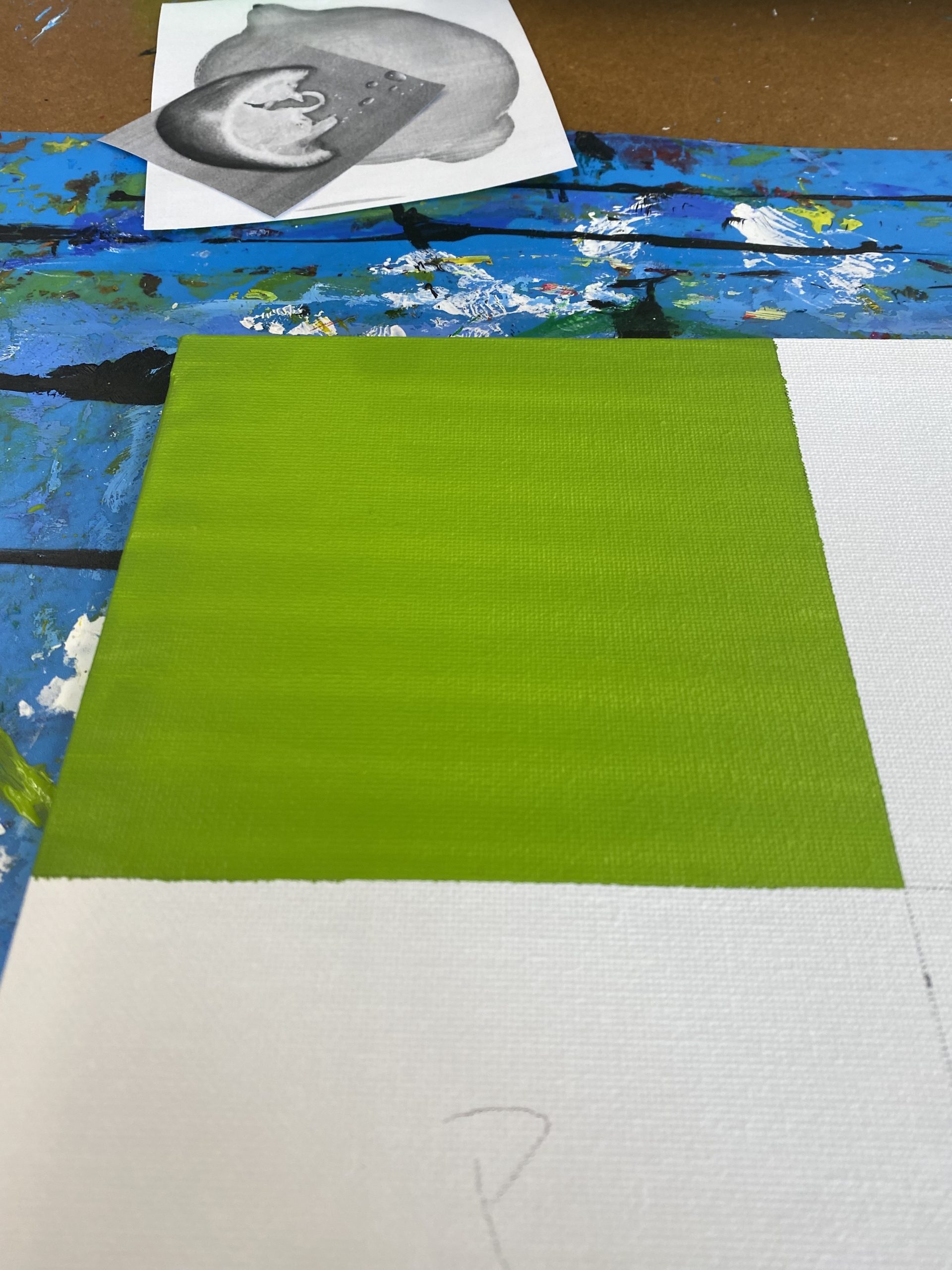 A rectangular canvas divided into four sections: the top left painted green, the other sections left unpainted white. The canvas rests on a paint-splattered blue surface.