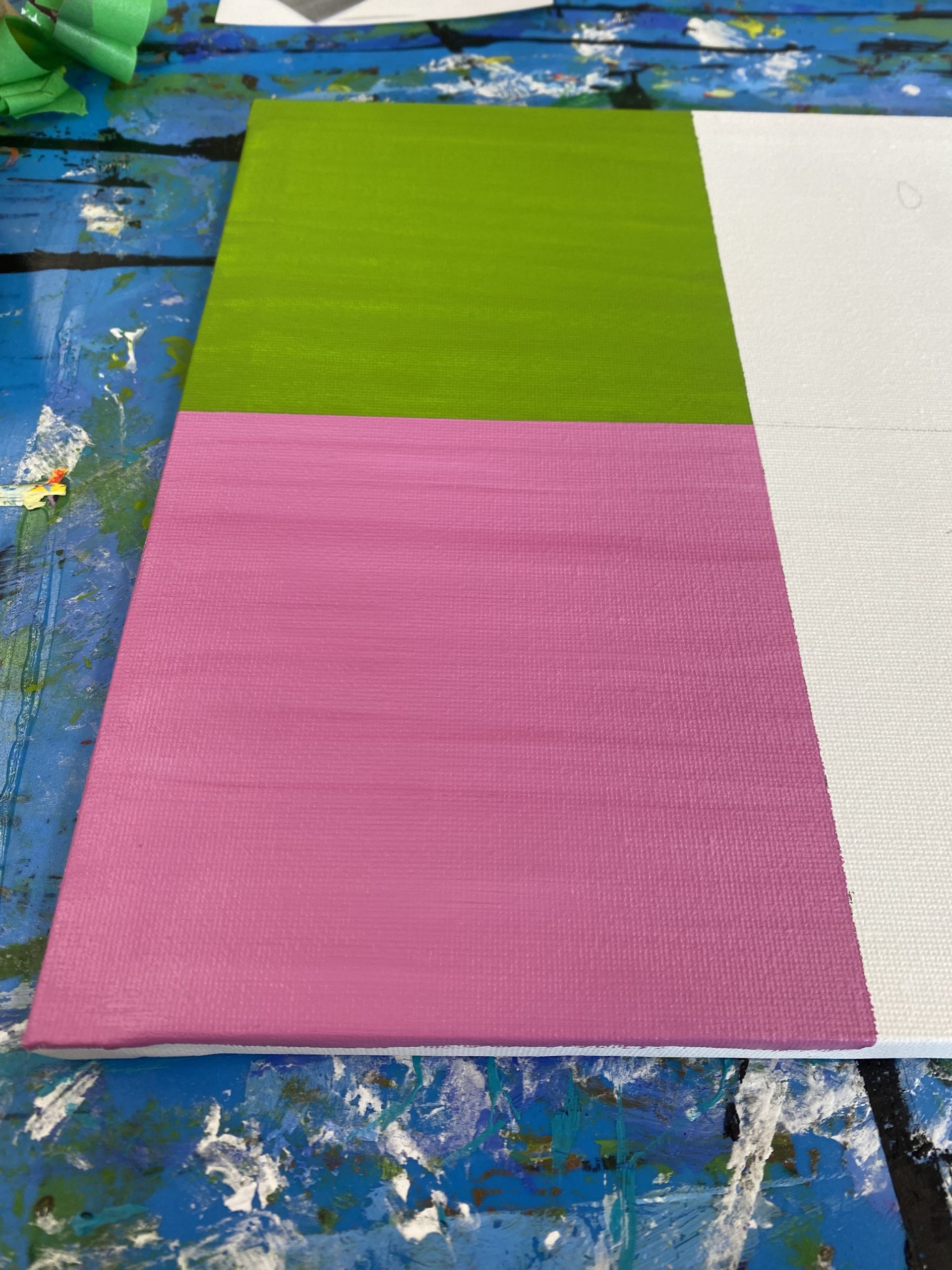 A rectangular canvas divided into four sections: the top left painted green, the bottom left painted pink, the other sections left unpainted white. The canvas rests on a paint-splattered blue surface.