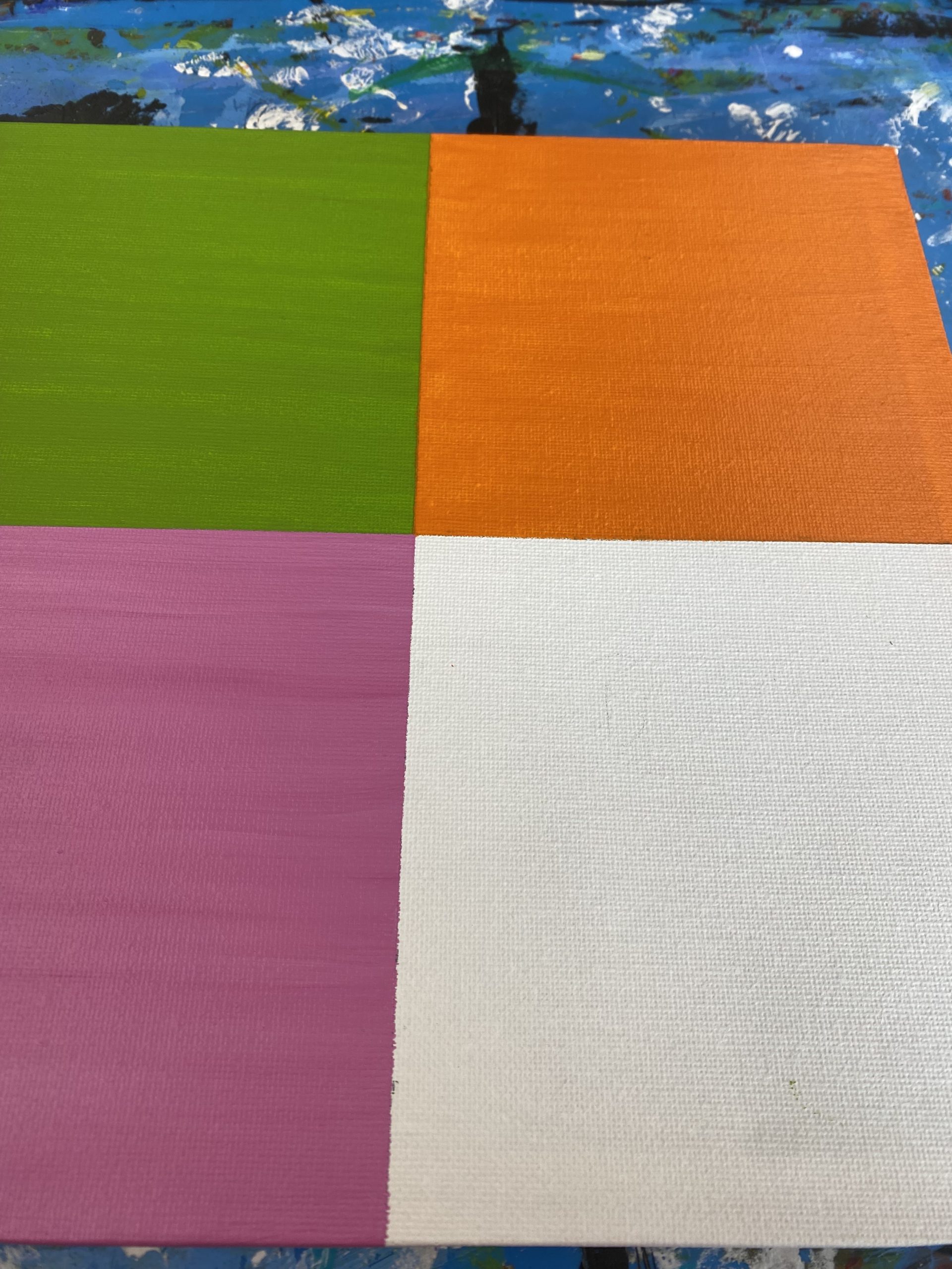 A rectangular canvas divided into four sections: the top left painted green, the bottom left painted pink, the top right painted orange, the other section left unpainted white. The canvas rests on a paint-splattered blue surface.