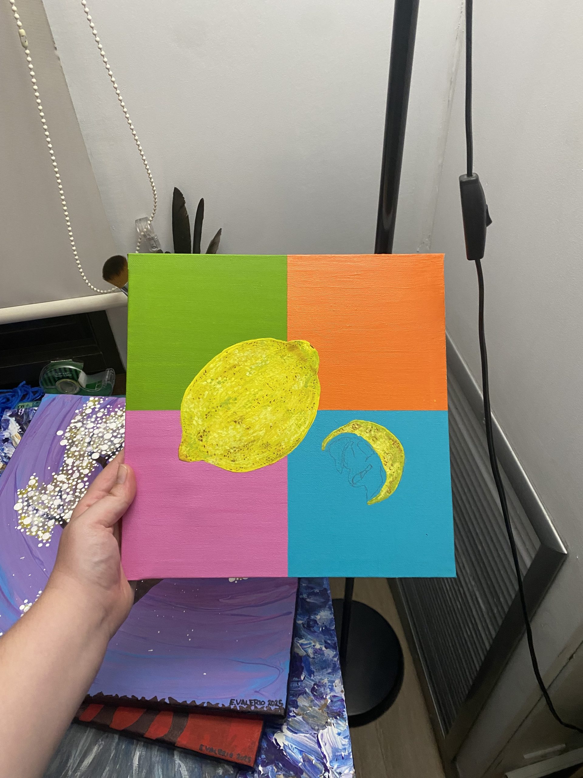 A hand holding a rectangular canvas divided into four sections: the top left painted green, the bottom left painted pink, the top right painted orange, the bottom right painted blue. On top, a yellow lemon and a yellow crescent-shaped lemon slice are painted.