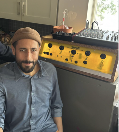 a picture of Carmen Papalia wearing a blue shirt a cotton cap and smiling through their tash. The Weather warlock sits behind them in their home space.