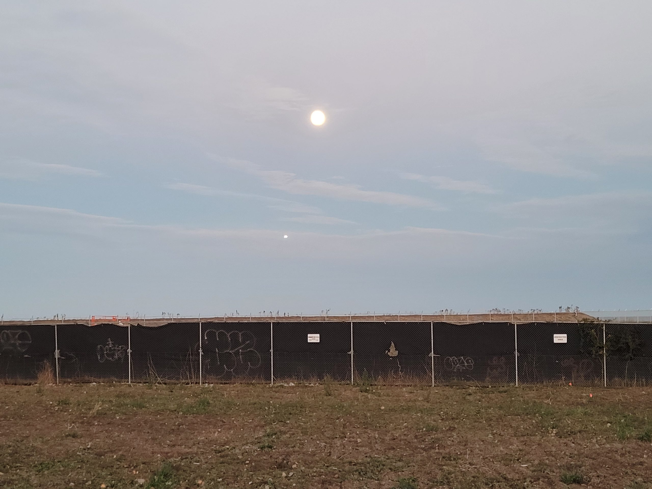 A photo of a large shy with the sun behind the clouds. In the foreground is a large black fence and scrubland