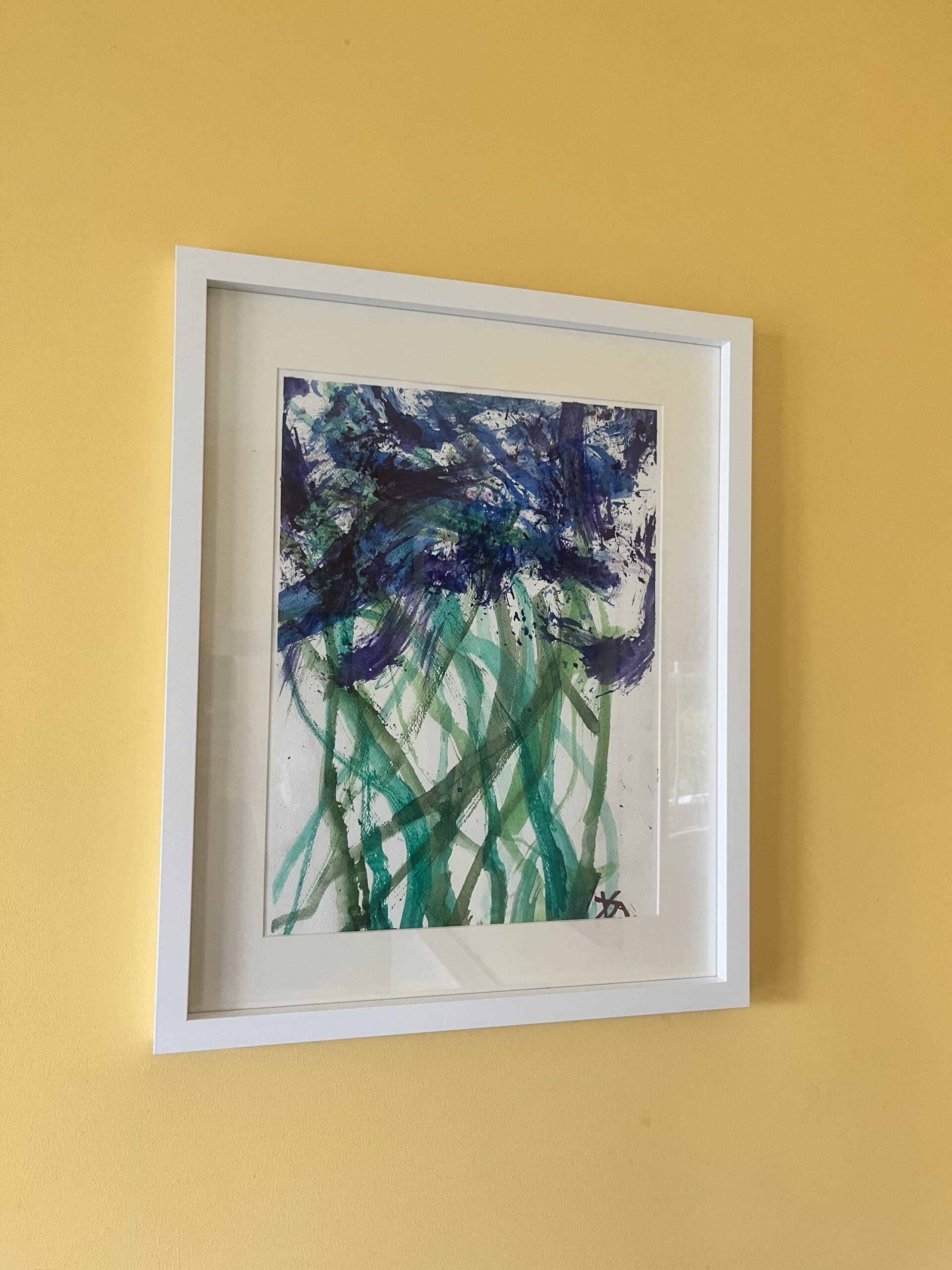 A framed painting of blue/purple flowers on different tones of green stems in an abstract style hanging on a yellow wall.