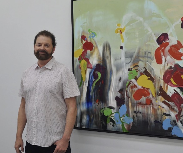 A white man with brown hair and facial hair stands next to a painting in a gallery. The painting is large, around twice the size of the man and uses bright flowers like splodges on a muted smudgy background. The background fades from darkest colours at the bottom to a greeny cream at the top.
