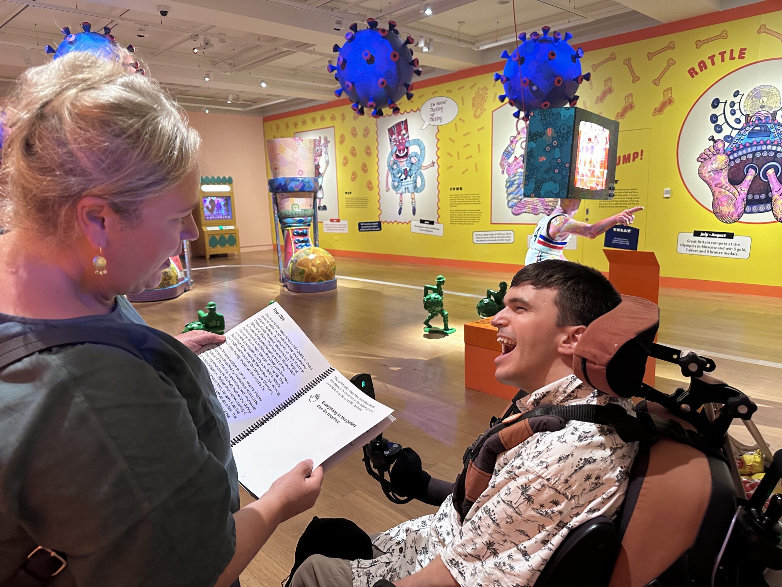 A white woman is holding a brochure and looking at it with a white man who uses a wheelchair. They are in a gallery space full of lots of different artwork both on the walls and on plinths in the middle of this space. The walls are yellow and peach and the artwork is very bright and colourful.