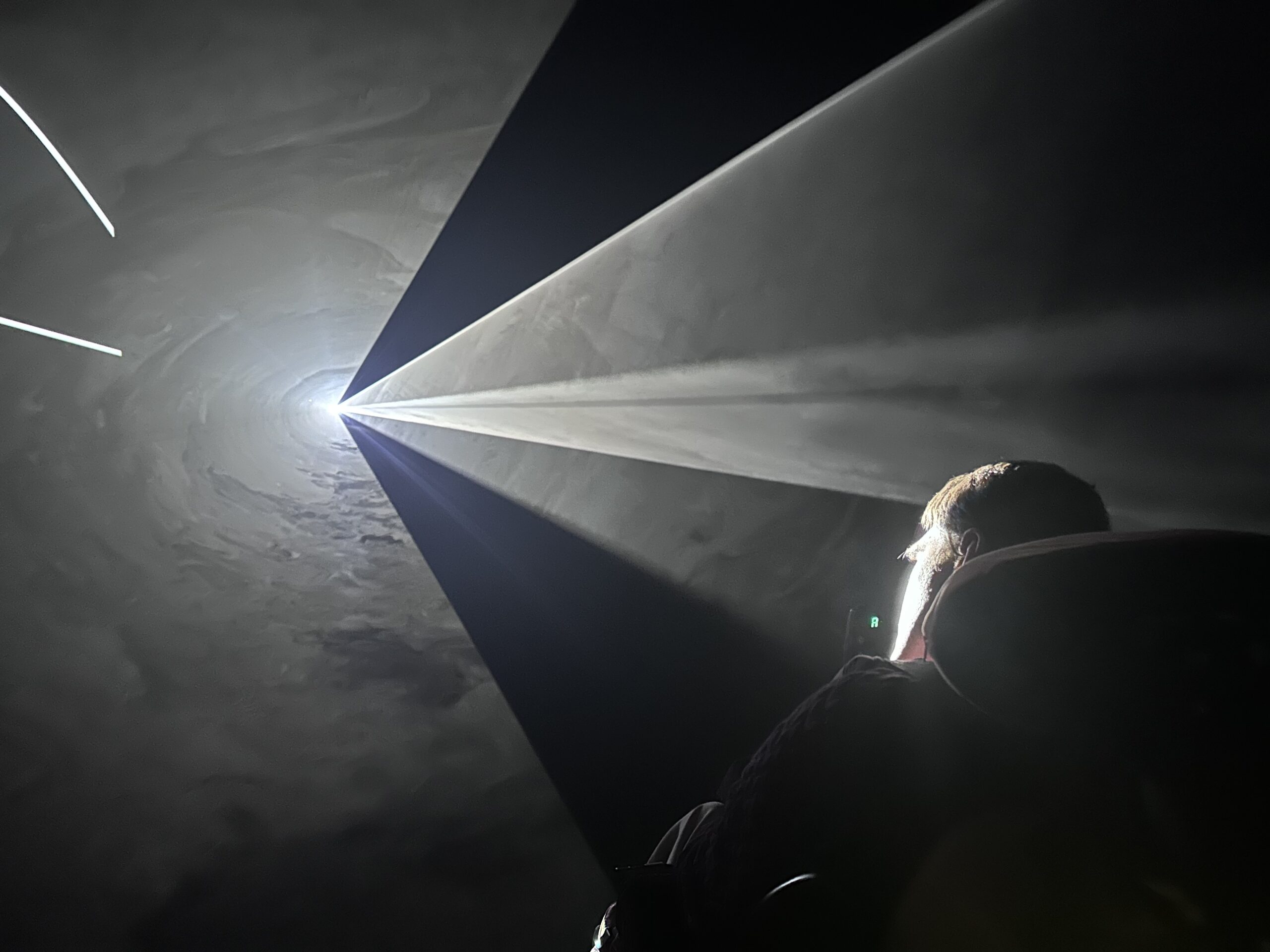 A man with dark hair looks to the left into light streaming in triangles. Everything is black and grey. apart from the man's face which is illuminated in the light to show his white skin and brown hair.