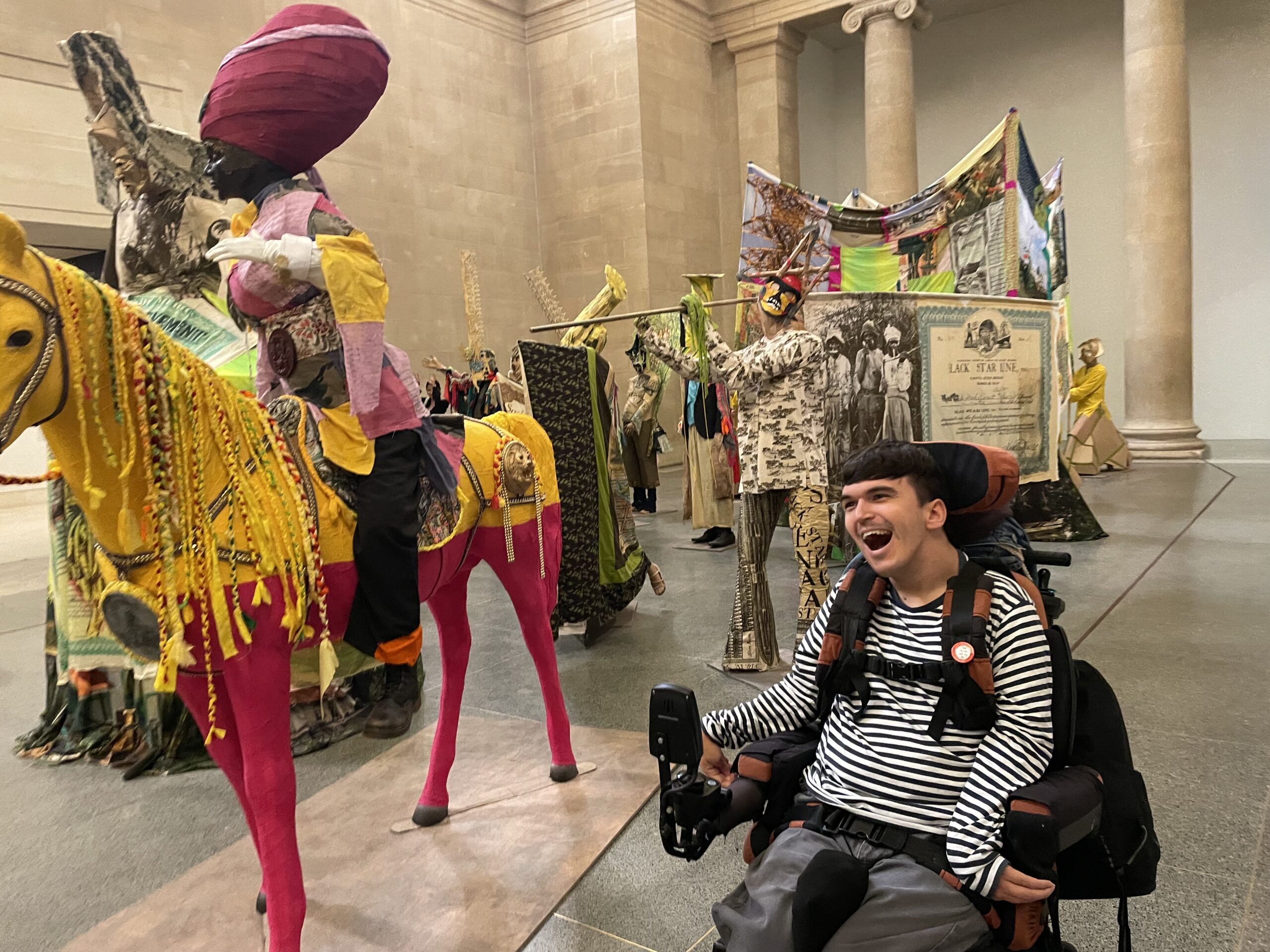 a young white man with dark hair and a striped top who uses a wheelchair is situated in front of large sculptures of a parade. The sculptures are colourful and made using a range of materials such as fabric and the man is smiling. There is a pink and yellow horse in the foreground.