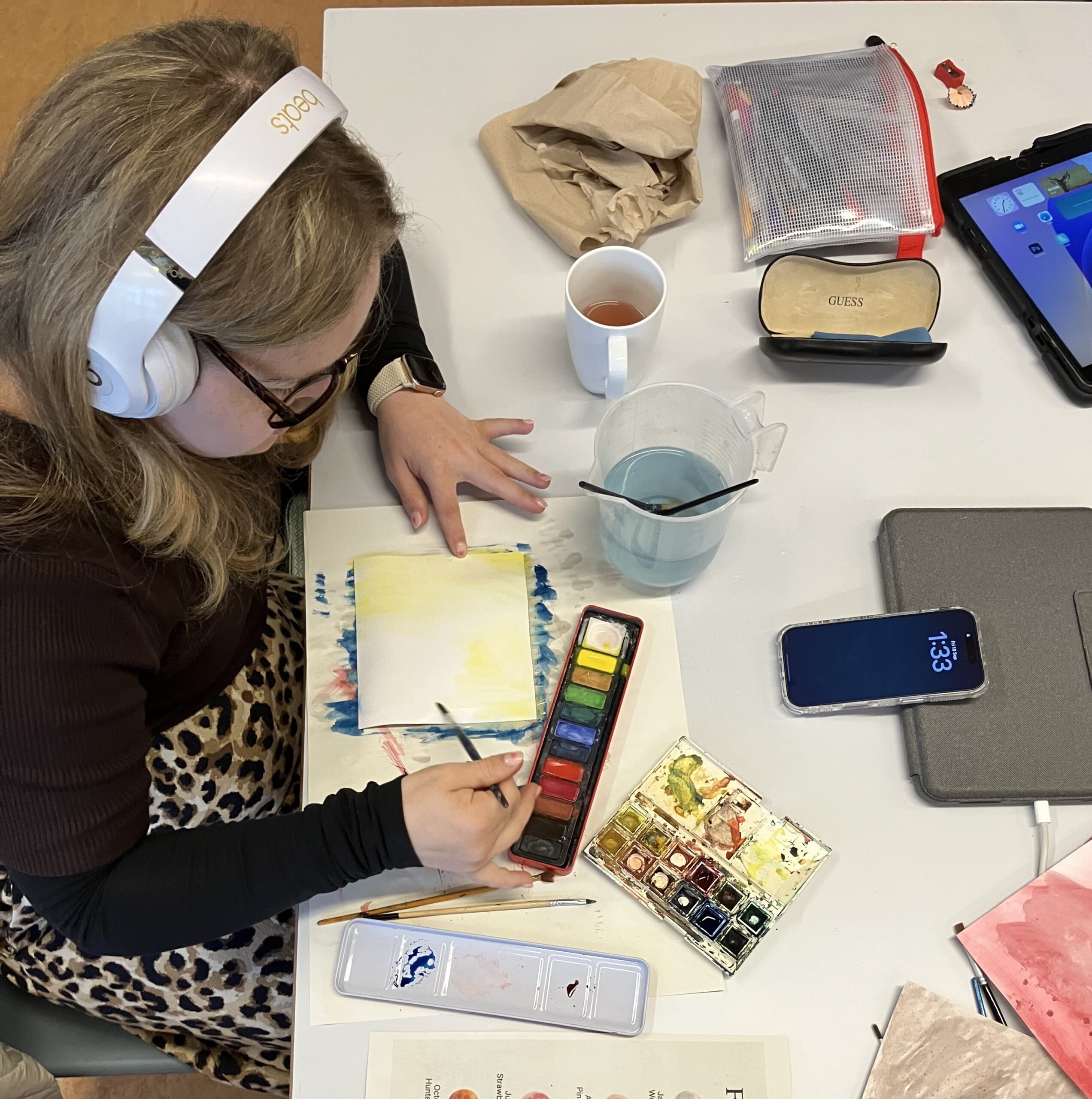 This photo shows Maddi sitting at her desk in the studio using watercolours. She has her watercolours, paint brushes, her phone for her music and her glasses case around her. She is wearing her headphones and painting yellow watercolours onto a square of paper.
