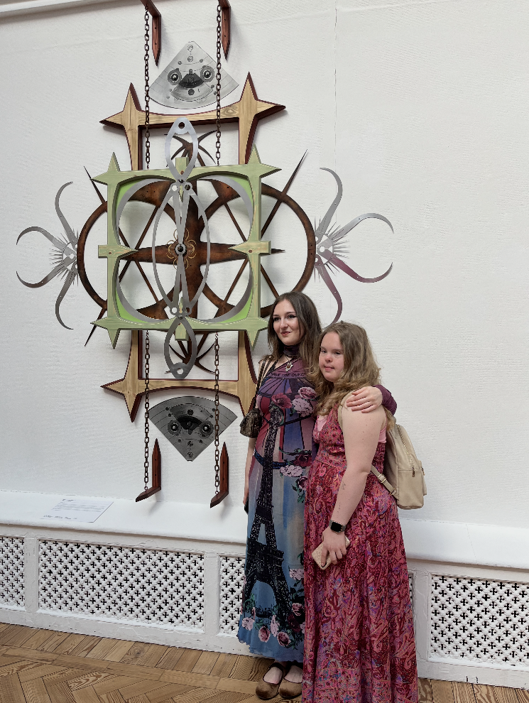 This photo shows Maddi and her sister Georgie standing in front of Georgie's art work. They are both wearing patterned dresses in similar colours of purple and red and black and blue. Georgie's dress has an image of the Eiffel Tower on the front. The art work is a sculpture hanging on a white wall. The sculpture is made of different materials and is in different colours of brown, green and silver.
