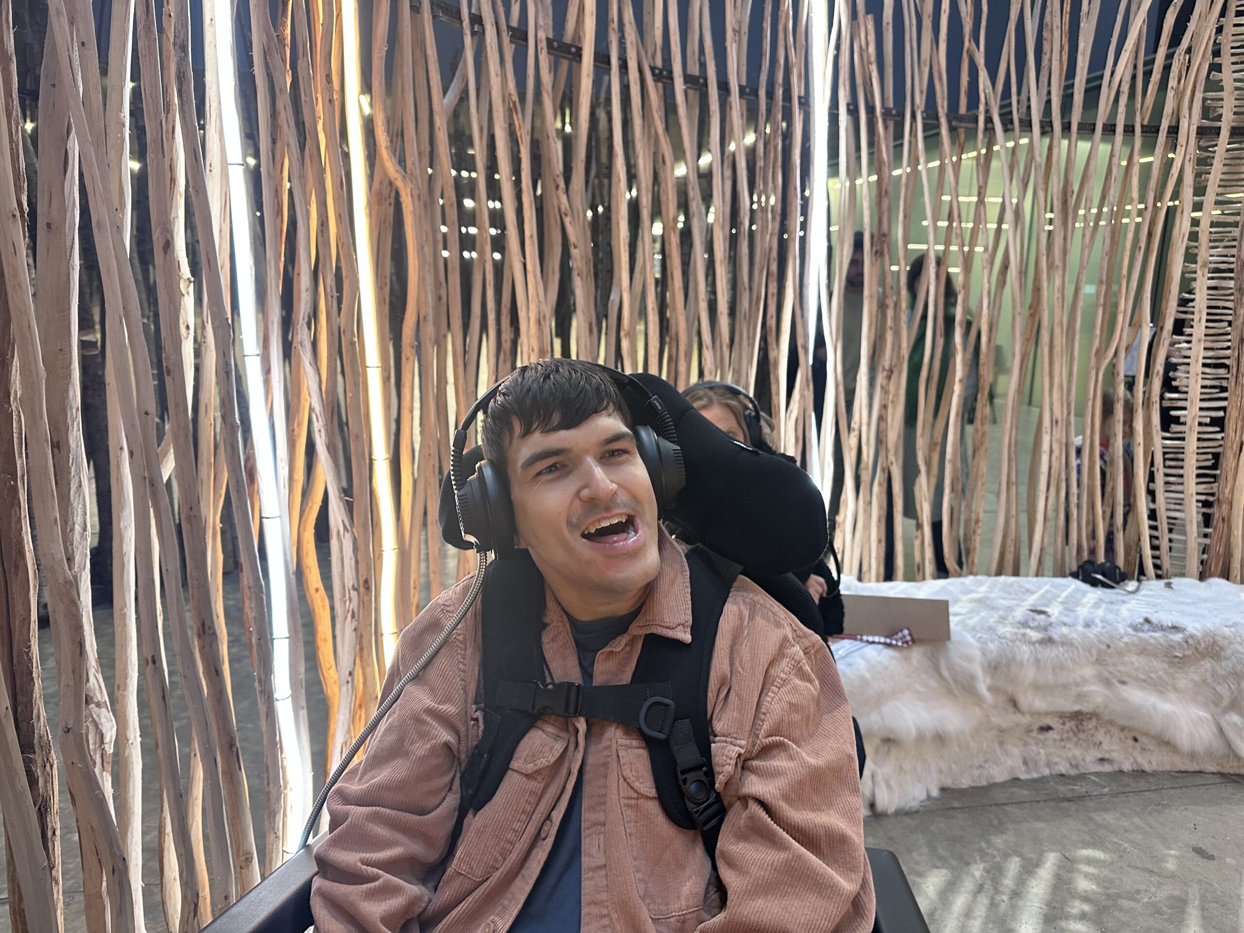 a photograph showing a young man in a wheelchair with dark hair looking happy. He's wearing headphones and a pink corduroy coat and is in a structure made from vertical sticks. to his right there is what looks like a bed covered with animal skins.