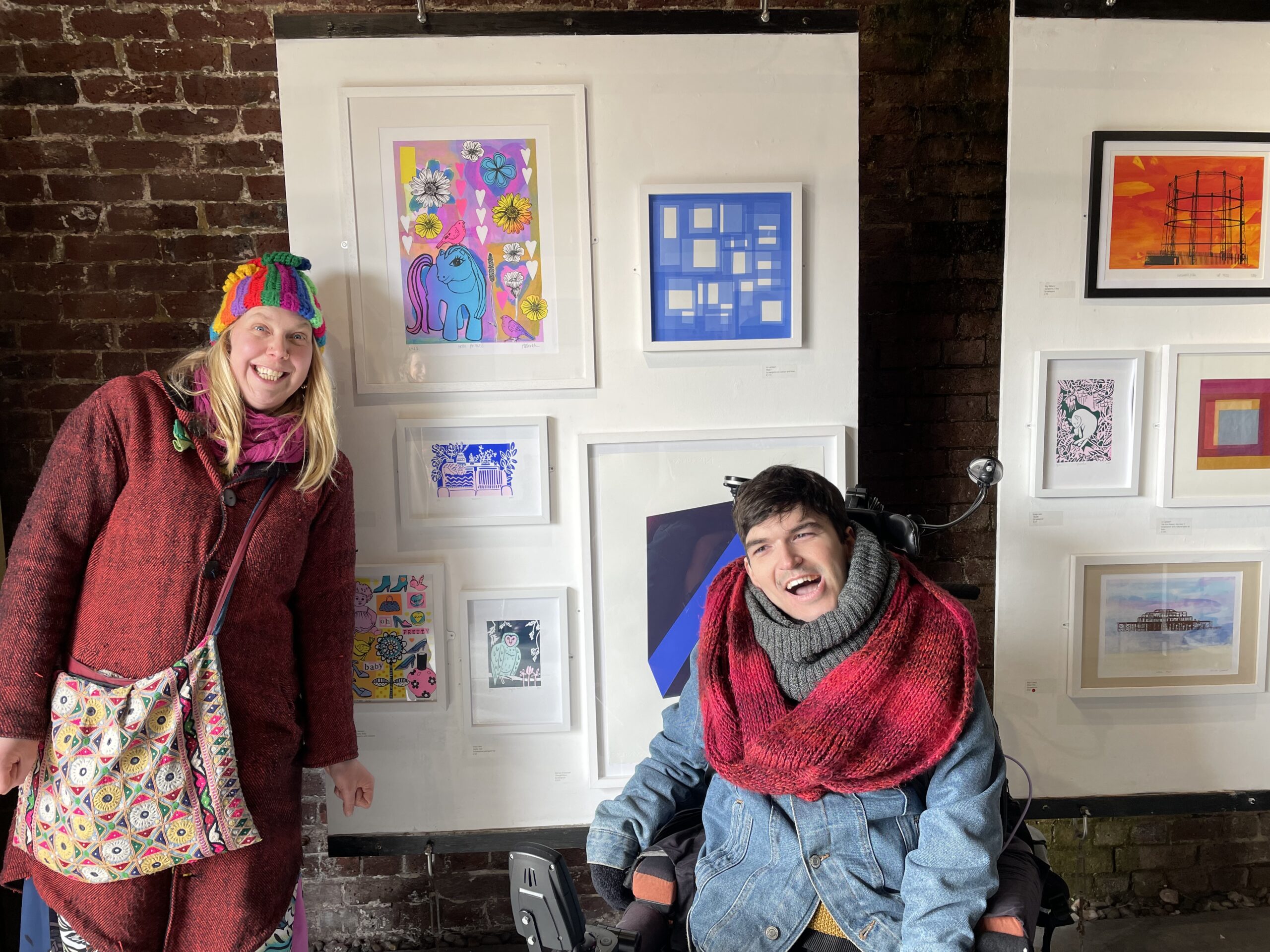 a white woman with long blonde hair and a colourful hat and a white young man with dark hair, a red scarf and using a wheelchair stand in front of some artwork. They are both smiling and looking happy. The artwork is in white frames and hung on a white panel. One of the images is a blue My Little Pony.