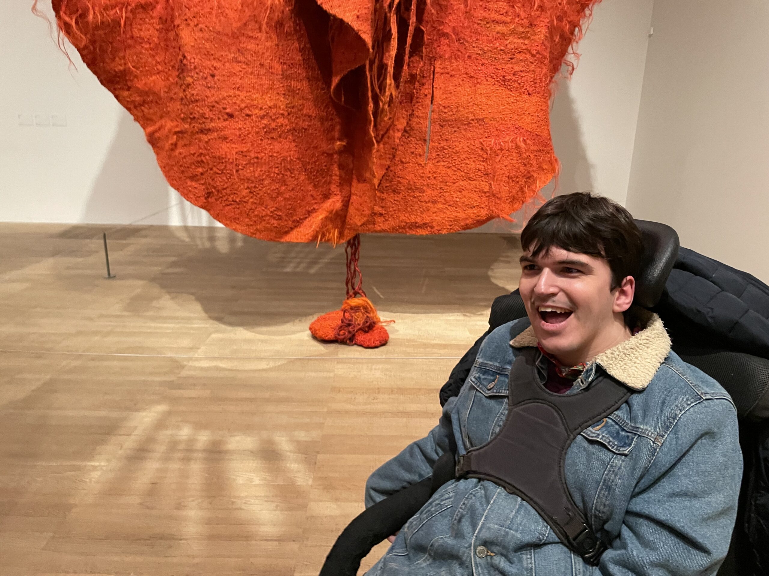 a young white man with dark hair who uses a wheelchair is smiling in the foreground. There is the bottom of a big artwork made from orange felt-like fabric in round shapes sewn together with some strands coming out of the bottom and sitting on the floor.