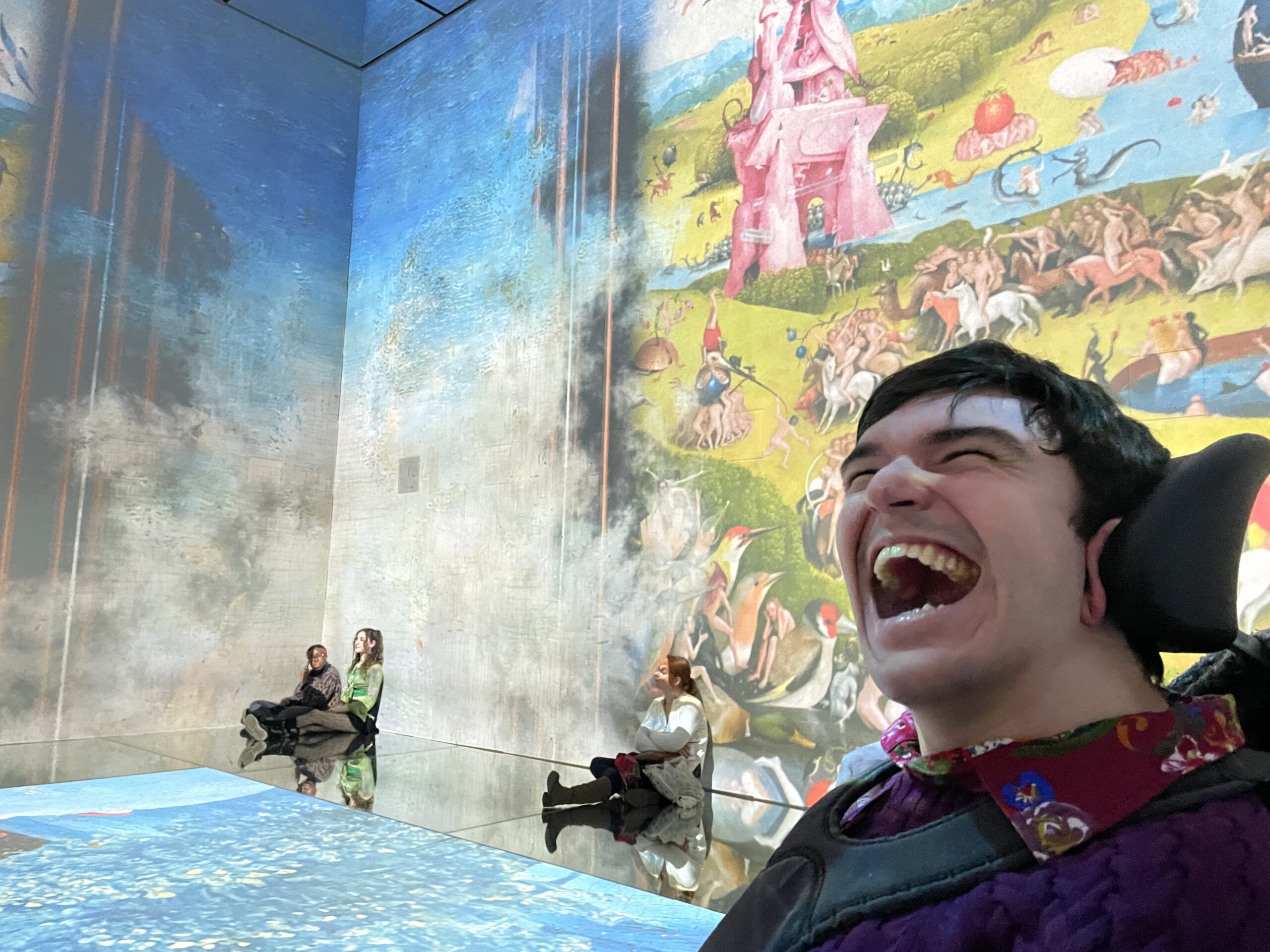 in the bottom right hand corner a young man makes a happy expression. He is white with dark hair and is wearing a red patterned shirt. Behind him shows the corner of a large room with artwork projected on the walls and floor. Some people are sitting on the floor looking at the art.