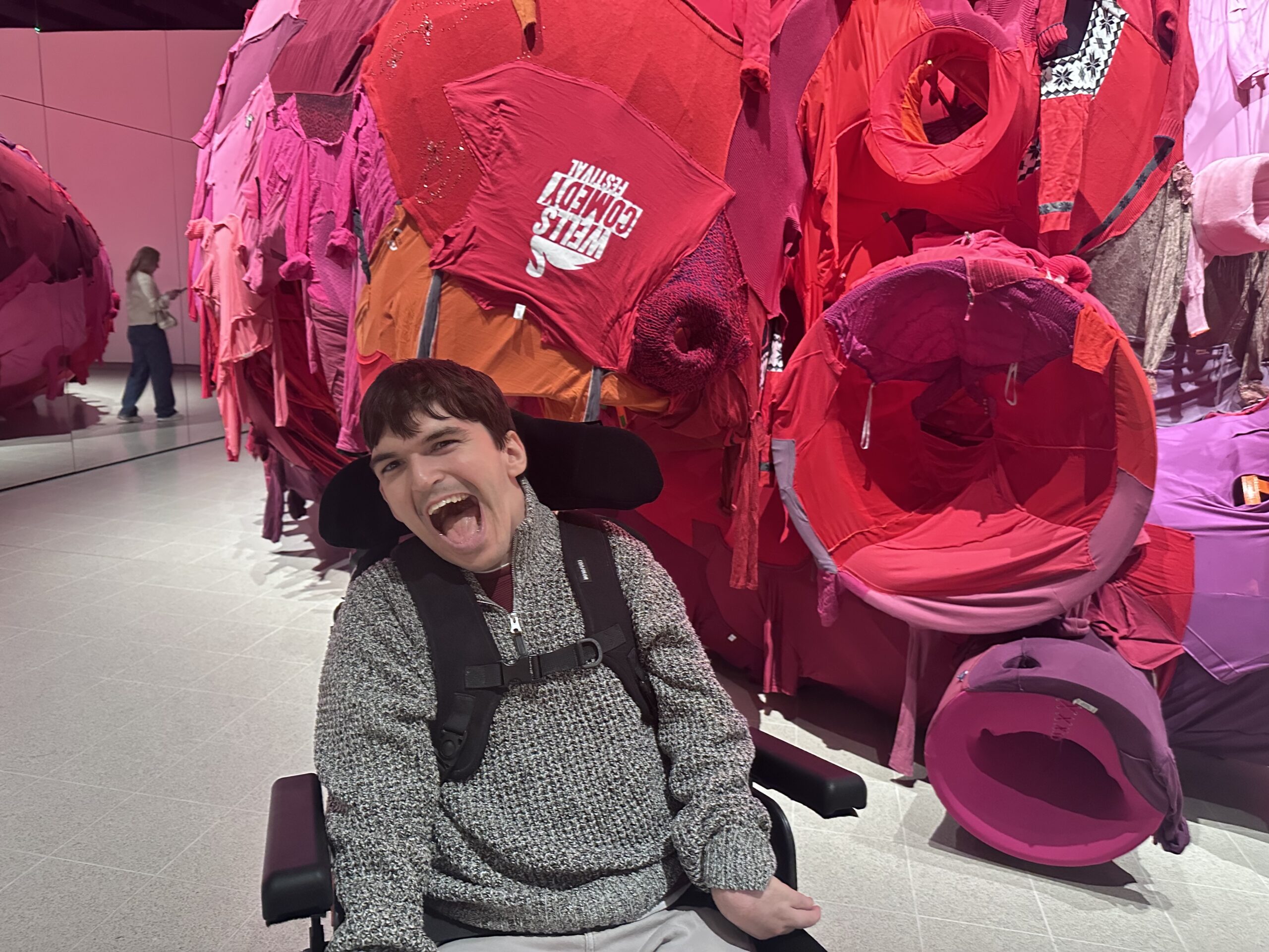a photograph showing a big bulging sculpture made from recycled pink and red clothes ‘Well comedy festival’ is printed on one of the T-shirts.there are circular shapes that the clothing is wrapped around. In front of the sculpture a young man who uses a wheelchair and has dark hair and white skin looks towards the camera with a joyful expression.