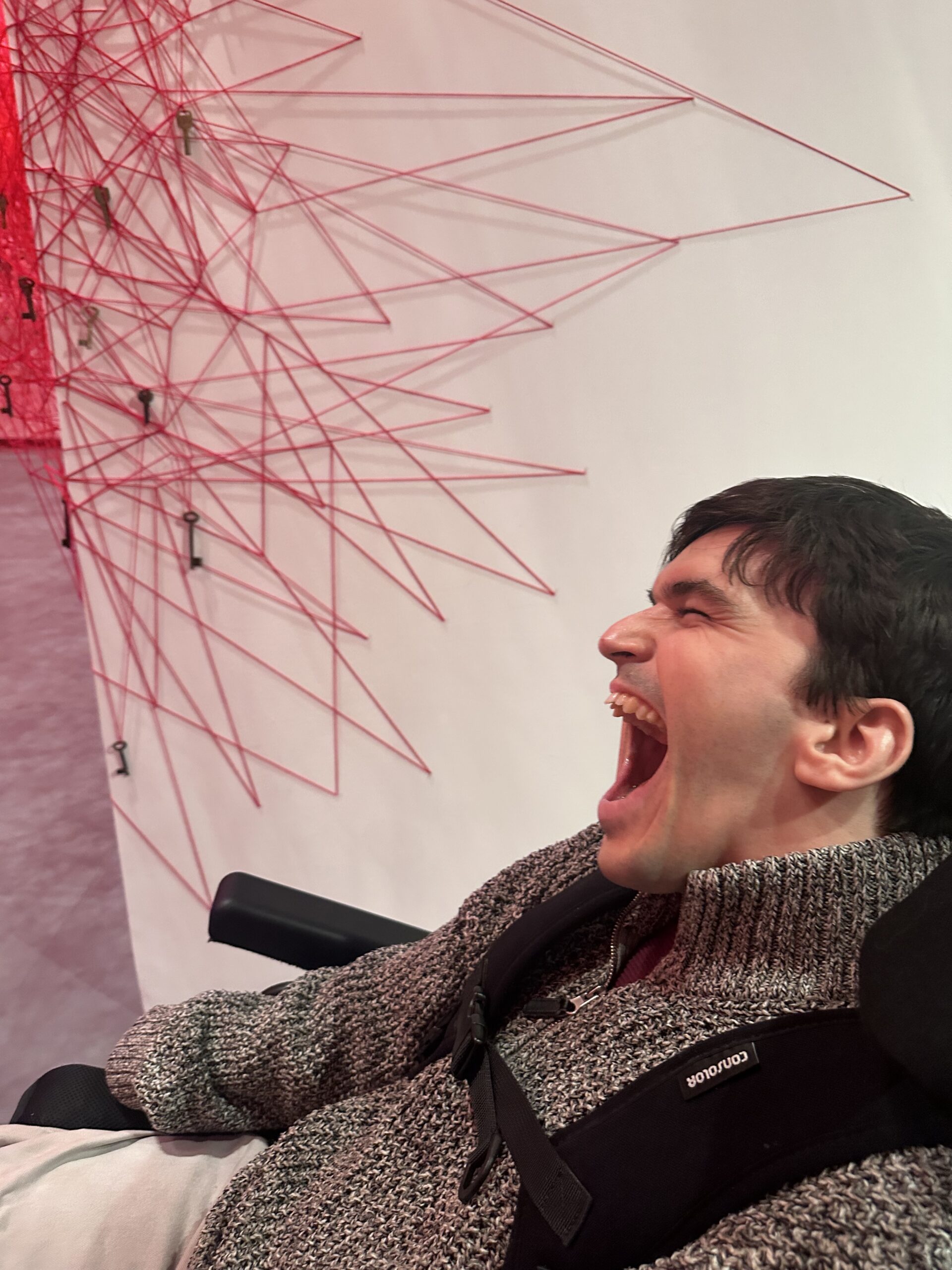 in the bottom right hand corner, a young man with dark hair and a grey jumper is making a happy expression. Behind him is a white wall with a crisscross of red threads creating a sculpture. keys hanging from the sculpture.