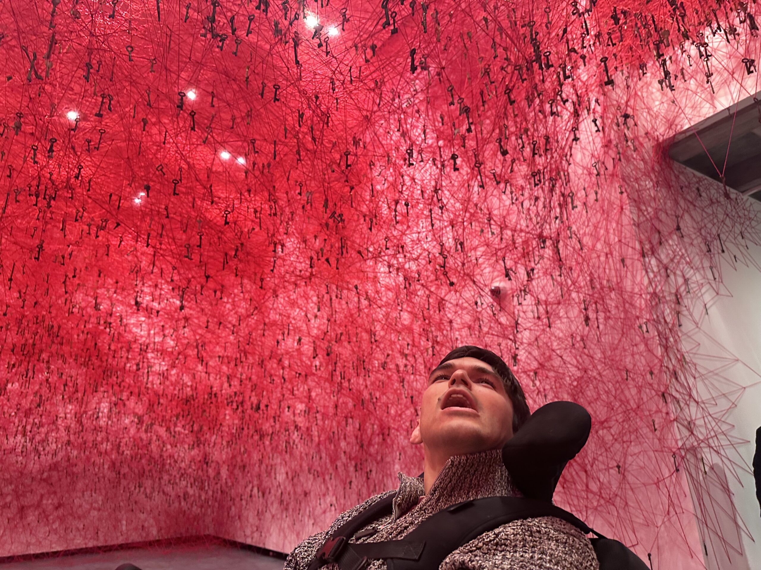 A photograph of a room where the walls are covered in inter-locking red threads creating a dome like structure keys hanging from the red thread. A young man is looking up at the sculpture.
