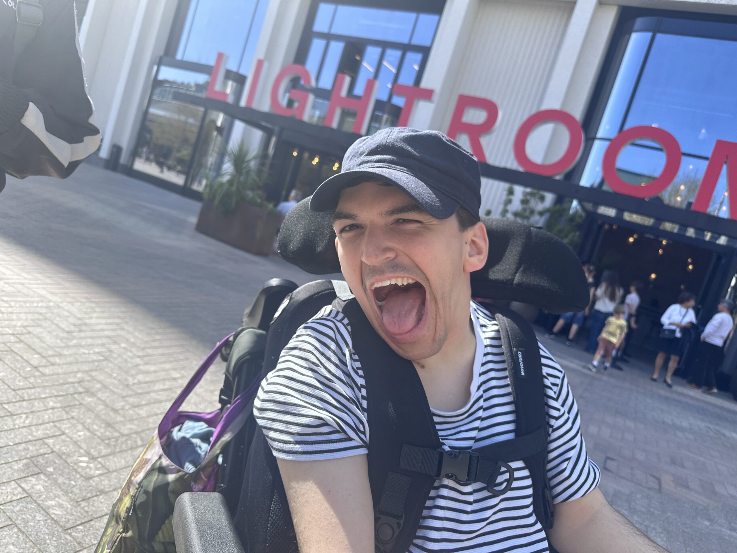 A photograph with the top half of a young white man's body who uses a wheelchair wearing a cap and looking happy behind him is a building with the words LIGHTROOM in large letters on the outside of the building.