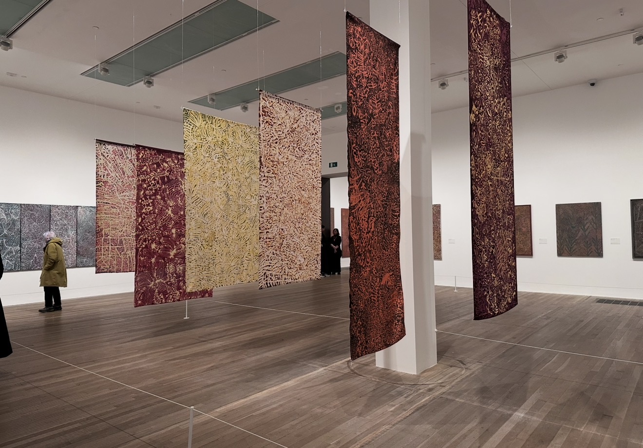 A photograph of lot‘s of long rectangular paintings hanging portrait from the ceiling in two rows one in front of the other in an exhibition space. There are more paintings on the white walls and people looking at the work. The paintings are two colours. One in the background and one on top. The top layer is abstract shapes and lines that together create a surface texture which spreads across the surface of the painting.