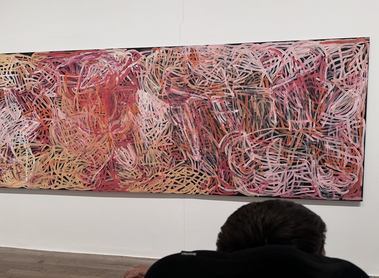 A large long rectangular canvas on a white wall. The top of a person's head and wheel chair in the bottom right corner. The painting has a black background with a mass of layers of orange, pink, red and yellow lines of similar thickness, weaving in and out of each other like a mass of wool.