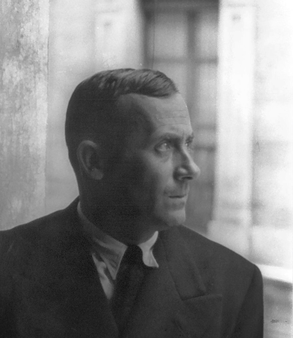 A black and white photograph of a white man with short hair looking to his left with a closed glass panelled door in the background.