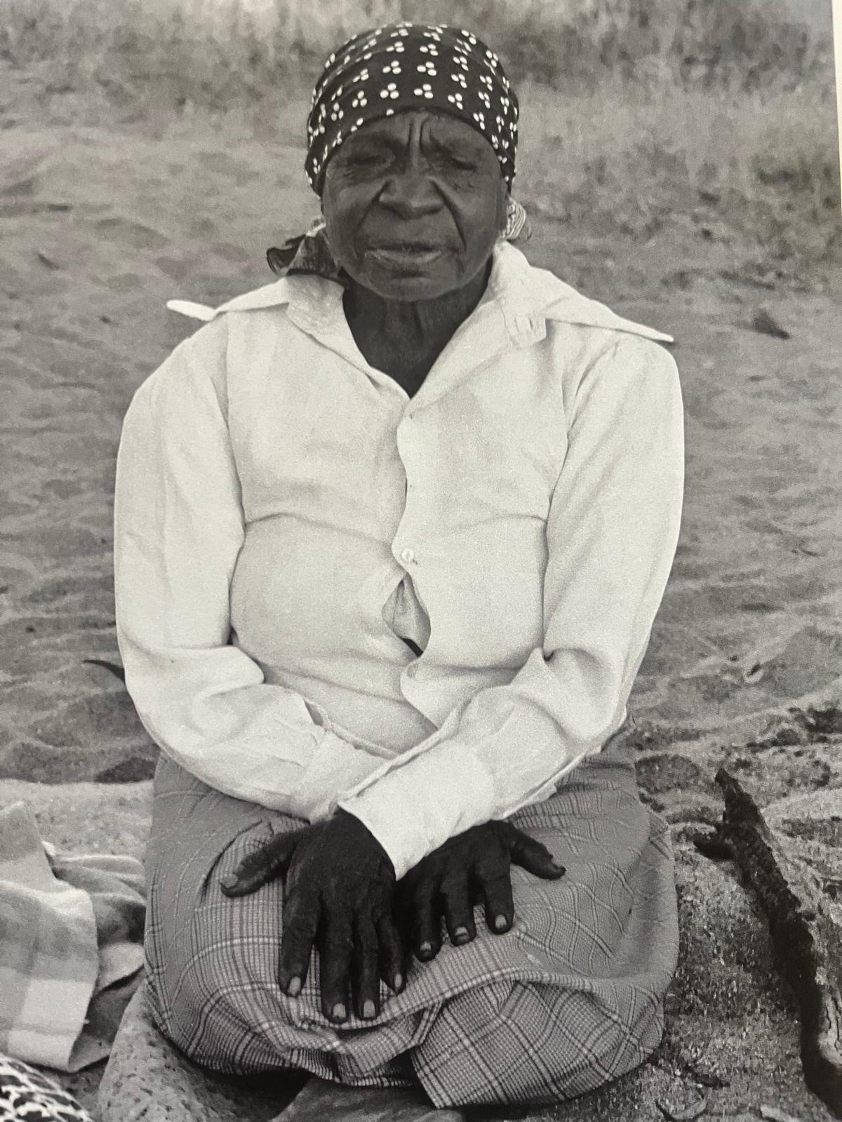A black and white photo of a black woman kneeling in the desert. She has her hands crossed on her lap and is wearing a head scarf.