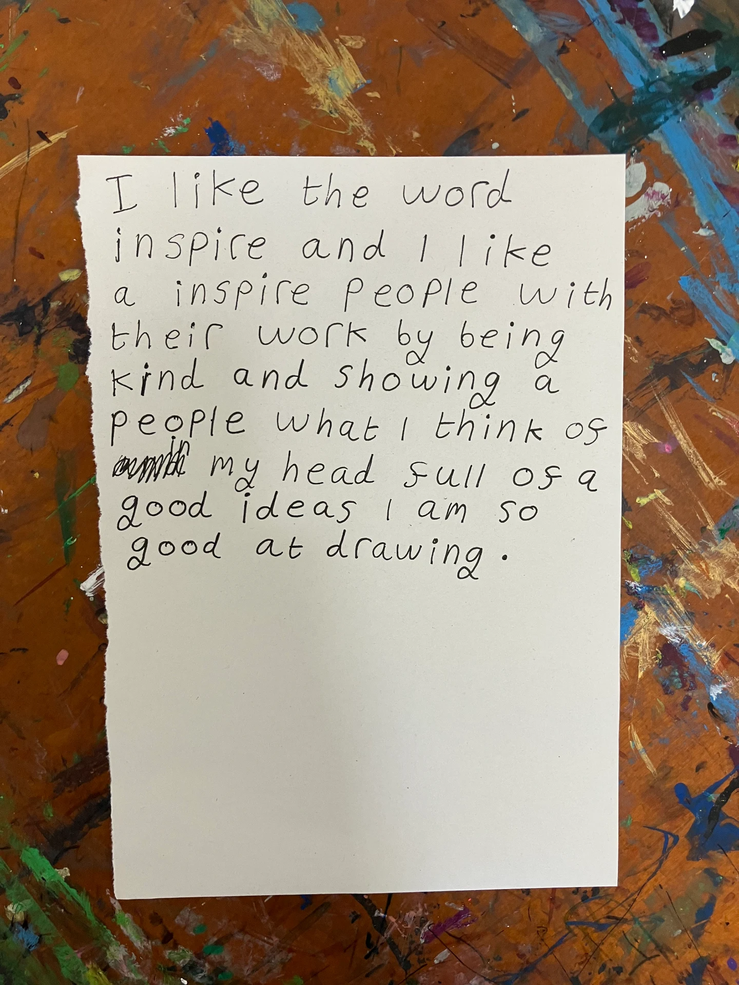 The photo is a close-up of a piece of paper resting on a paint-splattered brown table. The paint is yellow, blue, green, white and purple. The paper is white and in black pen Maddi has written the following: 'I like the word inspire and I like a inspire people with their work by being kind and showing a people what I think of in my head full of a good ideas I am so good at drawing.'