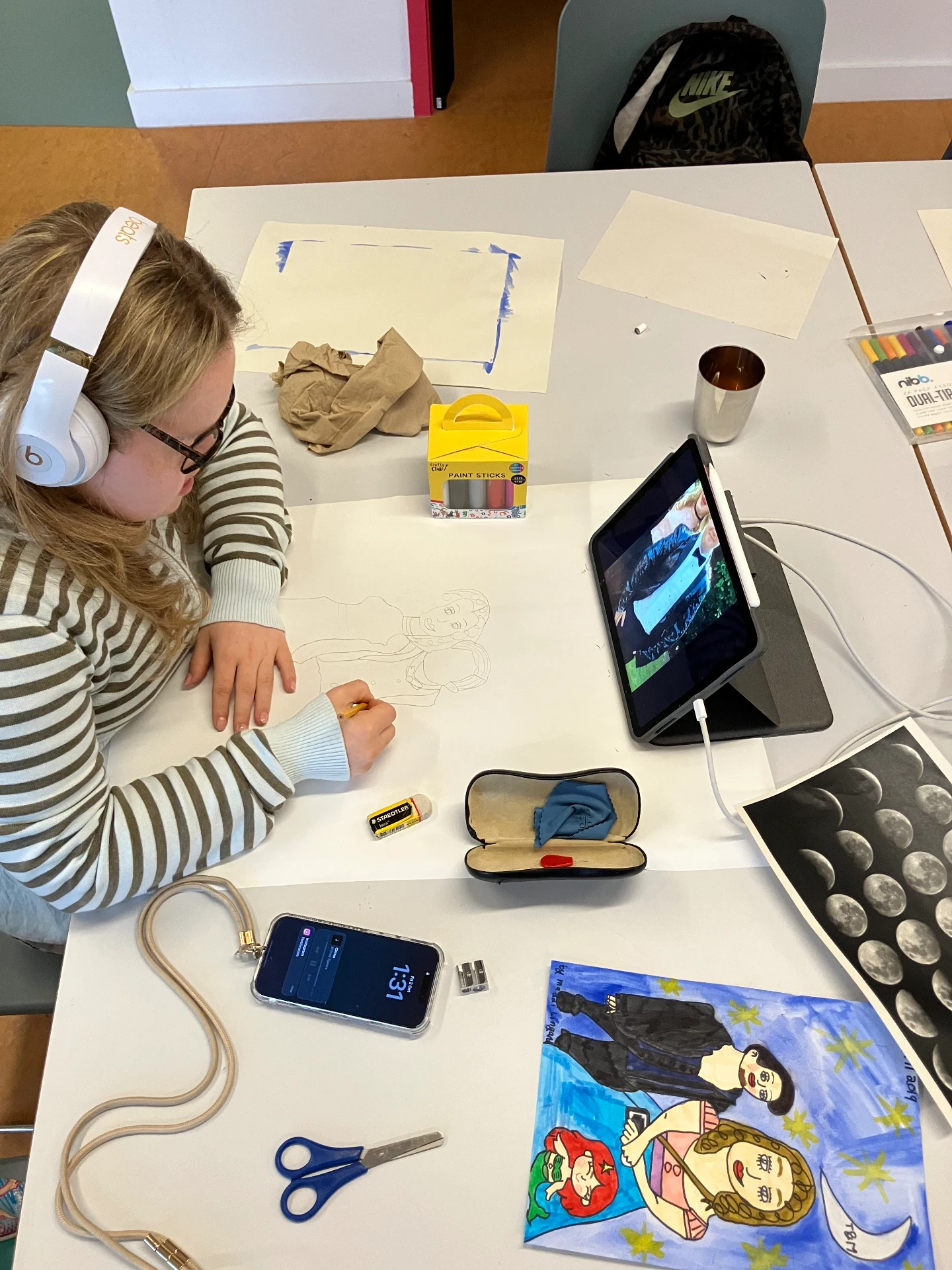 This is a photo of Maddi working at her desk wearing her headphones, she is sketching in pencil from a close-up of a photo on her iPad. Next to Maddi are her materials and her inspiration images. Maddi is wearing a stripy top.