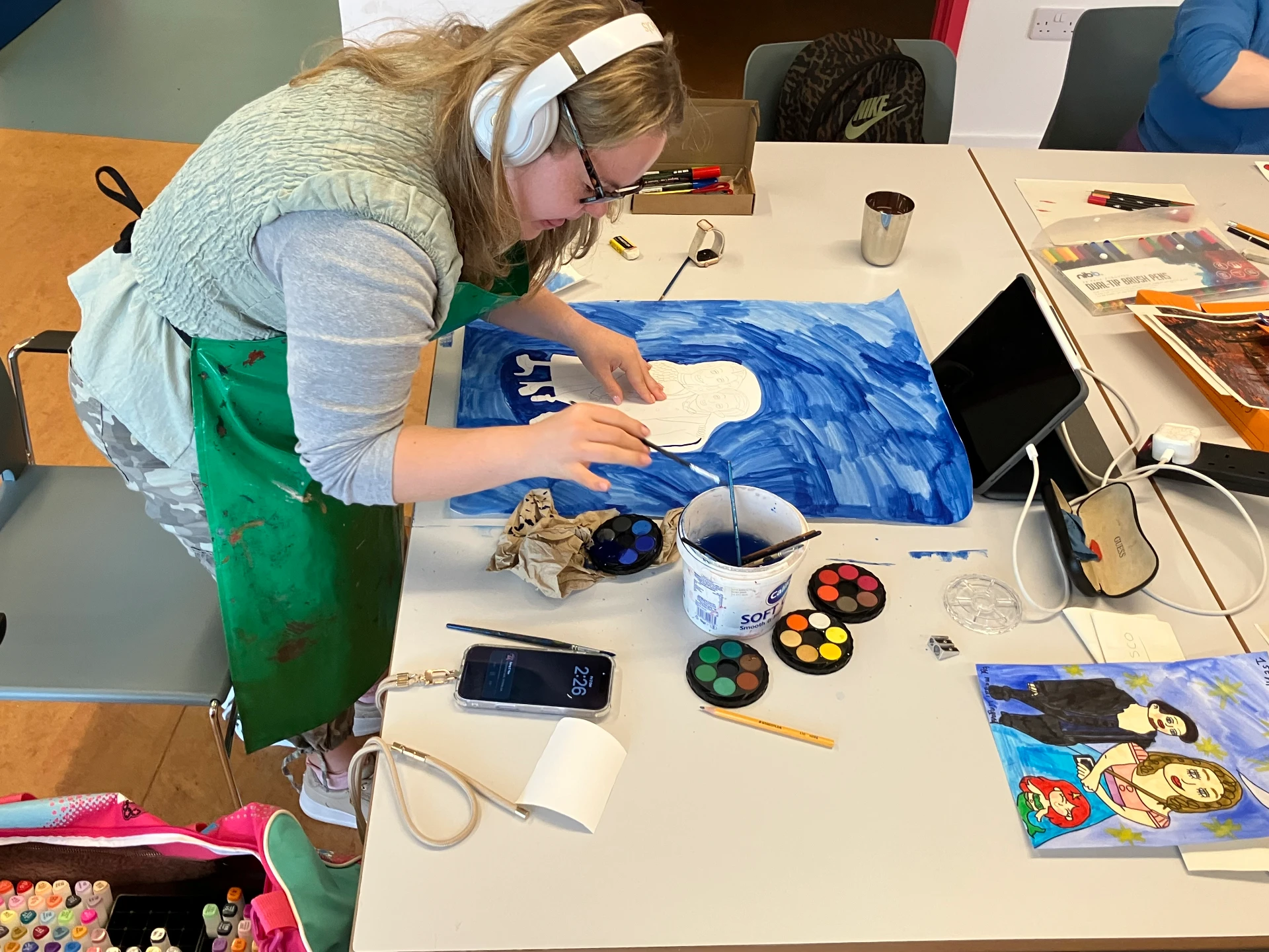 This is a photo of Maddi standing, working at her desk wearing her headphones, she is painting in watercolours. Next to Maddi are her materials and her inspiration images. Maddi is wearing a stripy top and a green apron.