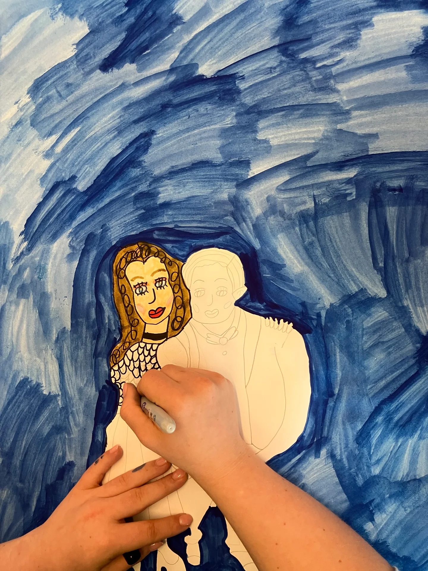 This is a close-up of Maddi's hands, she is working on a test drawing for her final work. It shows a pencil drawing of Maddi and her boyfriend Thomas. Maddi is adding black sharpie and felt-tip colour to her drawing. Surrounding the drawing is a textured watercolour sky in a dark and light blue colour.