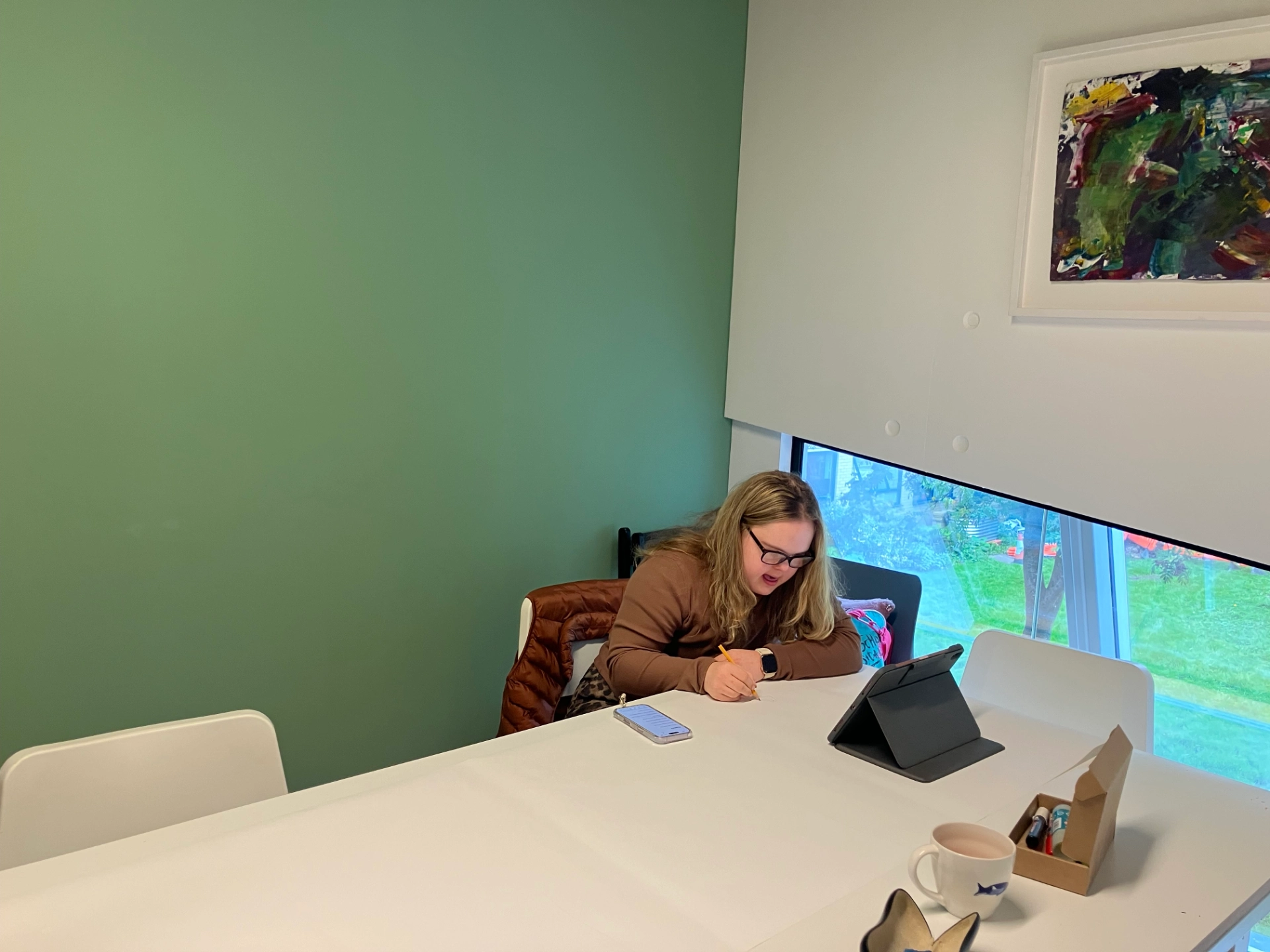 This photon shows Maddi sitting at the edge of a big white desk. The wall behind Maddi is painted dark green. Maddi is wearing a brown top and is drawing on a long sheet of paper, land has her iPad and drawing materials in front of her.