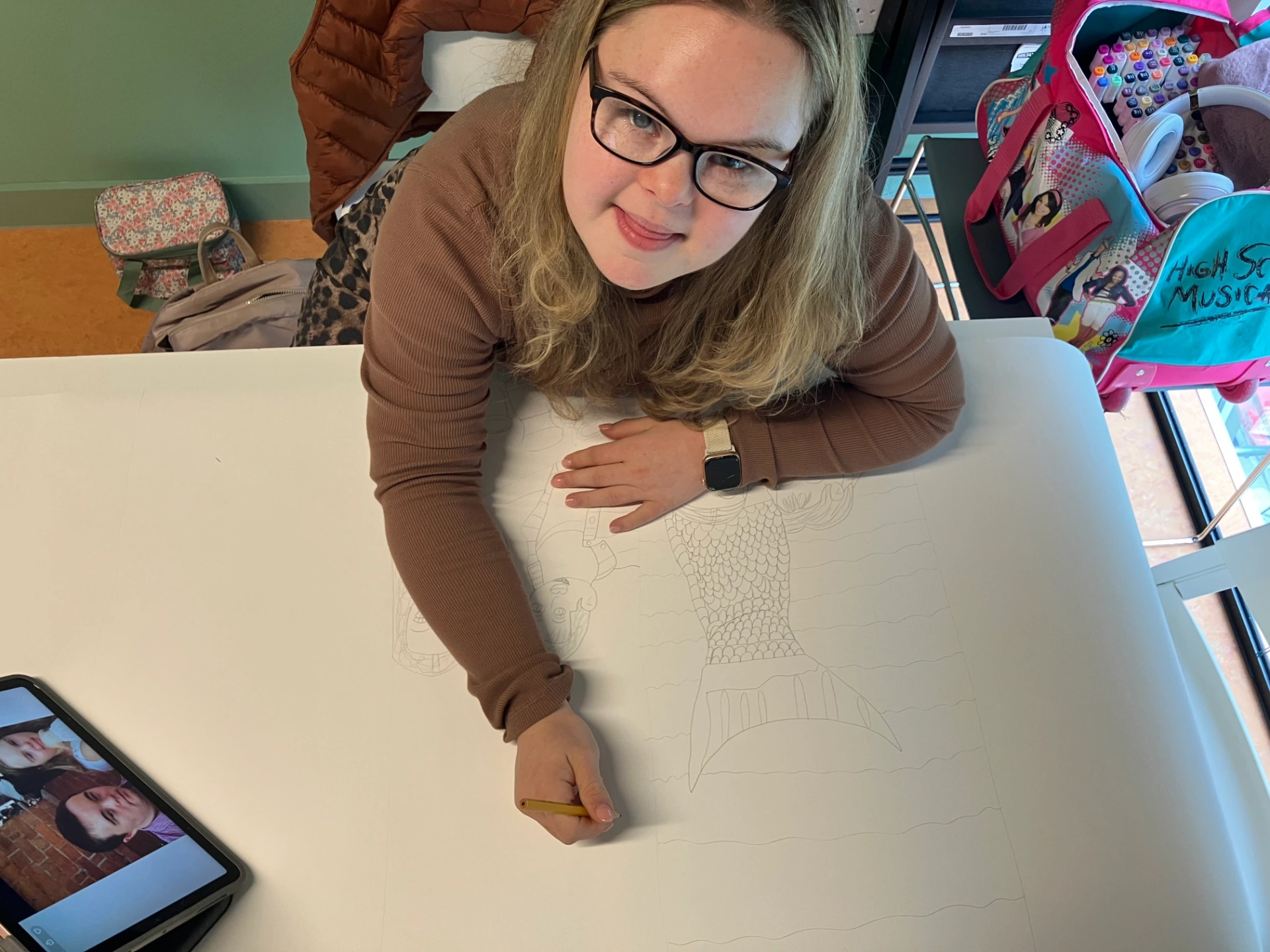 The photo is a close up of Maddi sitting at her desk in front of her work. The work is a pencil drawing on a large piece of paper. Maddi is wearing a brown top and glasses. The drawing is of Maddi wearing a mermaid costume, next another drawing of Maddi and her boyfriend Thomas.