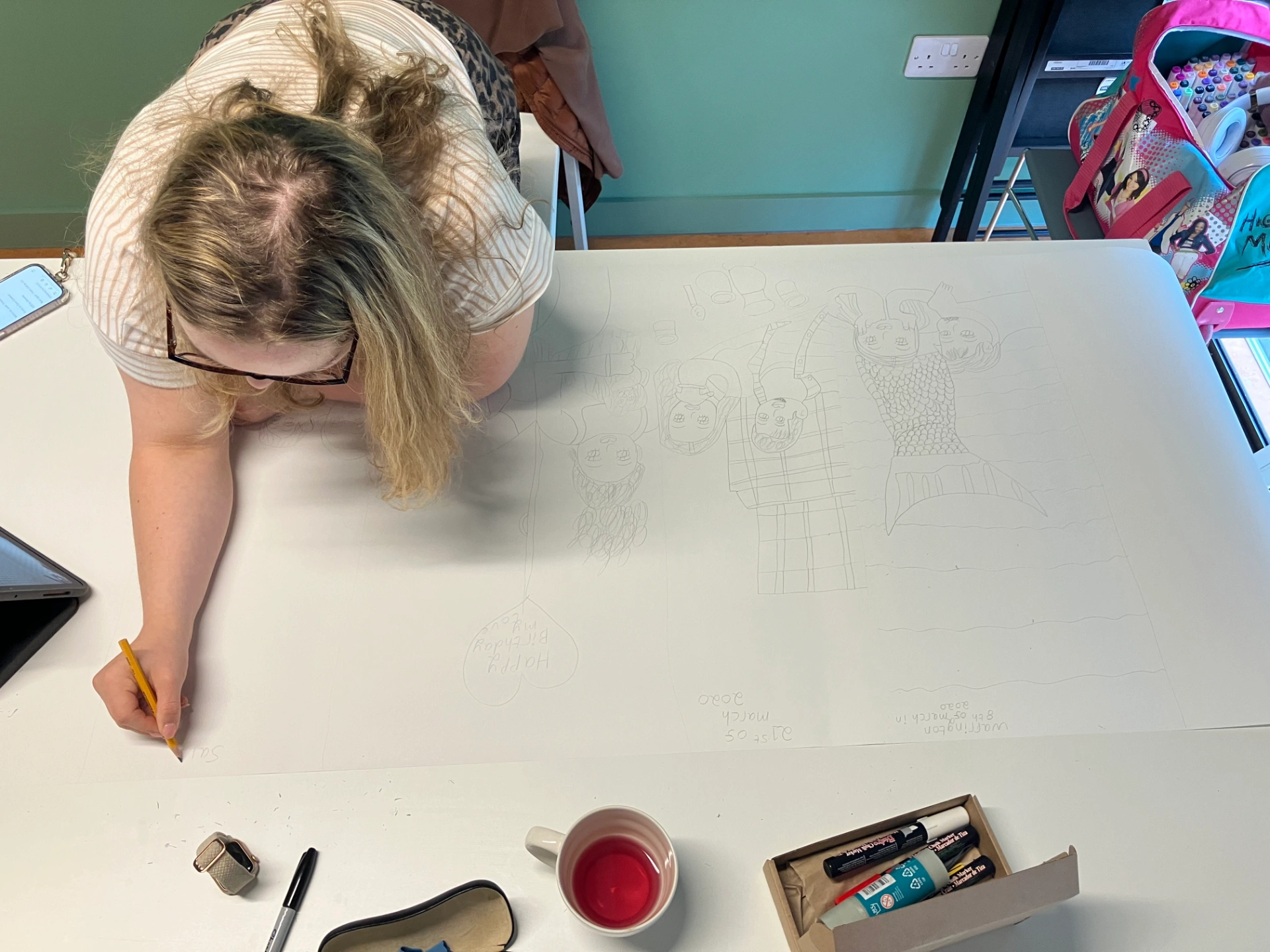 This photo is of Maddi standing, working at her desk on a large roll of paper. Maddi is drawing self-portraits in pencil. In front of Maddi are her drawing materials and mug.
