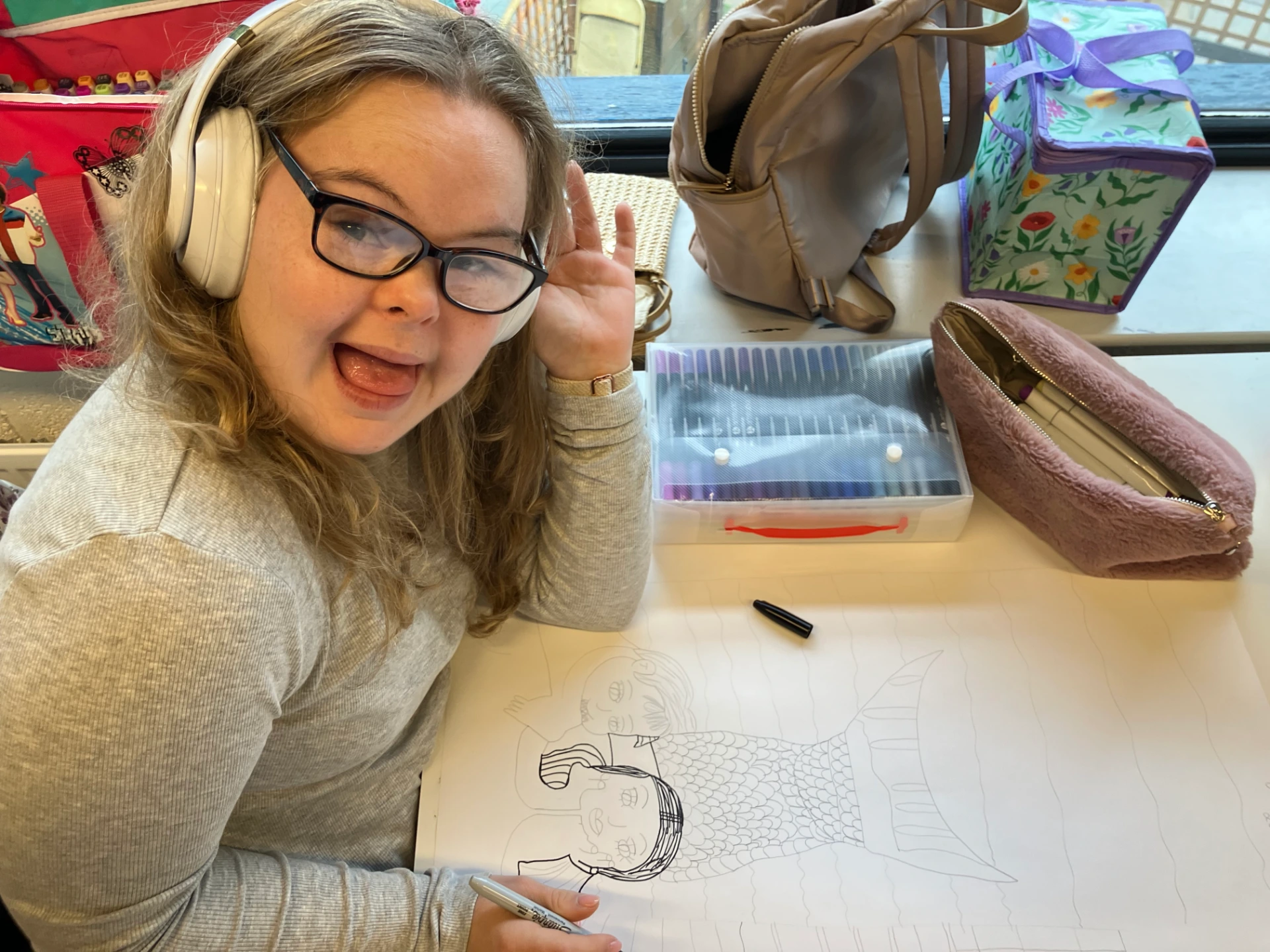 In the photo Maddi is sitting at her desk, wearing her white headphones and a grey top and glasses. She is on the left hand side of the frame. In front of her are her materials and her drawing on a large piece of white paper. Maddi is adding an outline in black sharpie pen. Maddi is looking and smiling and waving at the camera.