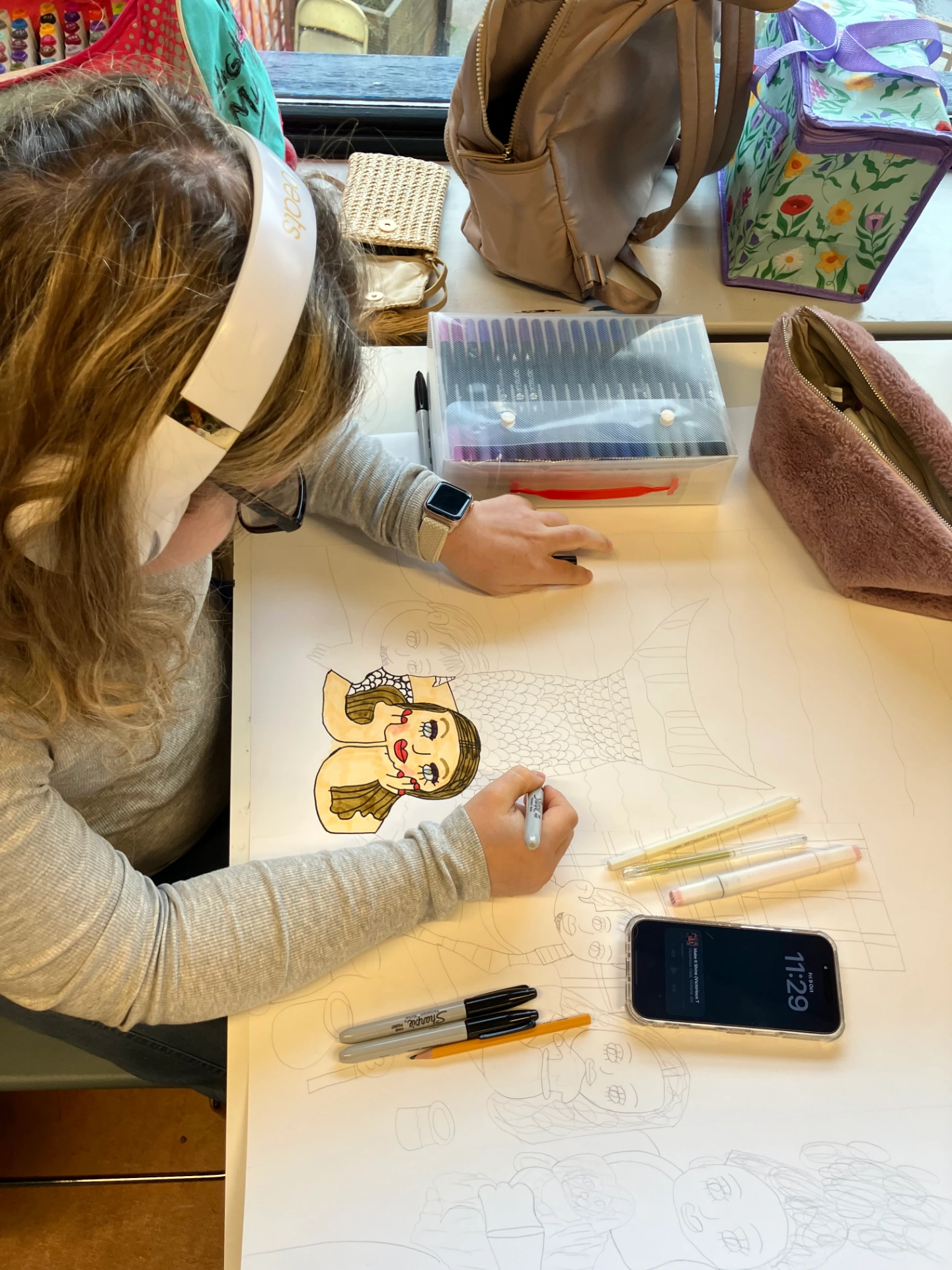 In the photo Maddi is sitting at her desk, wearing her white headphones and a grey top and glasses. She is on the left hand side of the frame. In front of her are her materials and her drawing on a large piece of white paper. Maddi is adding colour to her drawings.