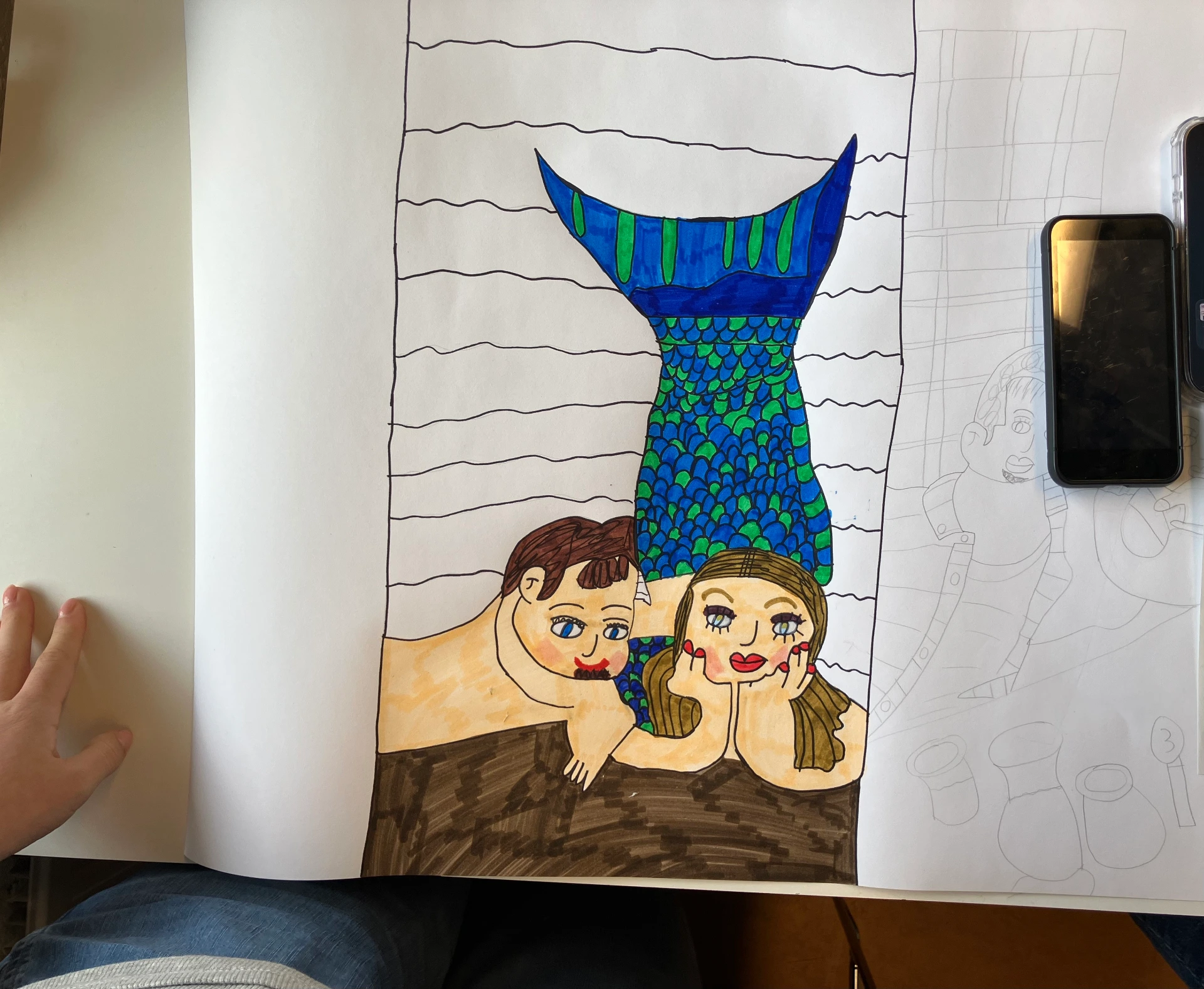 This image shows a close-up detail of Maddi's work. This is a drawing Maddi made of herself as a mermaid next to her boyfriend Thomas. The background is unfinished but Maddi's mermaid has a green and blue tail and blonde hair. They are both lying down on a brown surface.