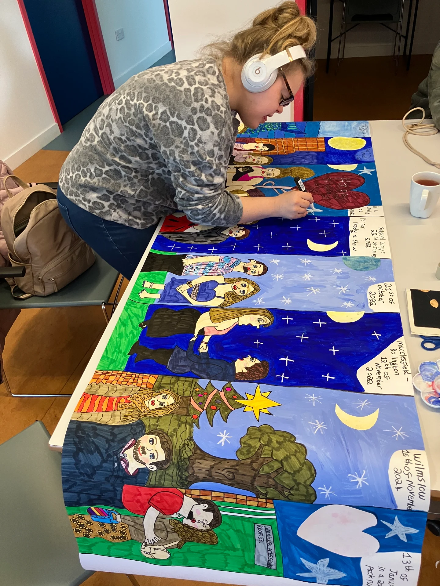 A woman wearing headphones leans over a long, colorful mural on a table, adding details with a marker.