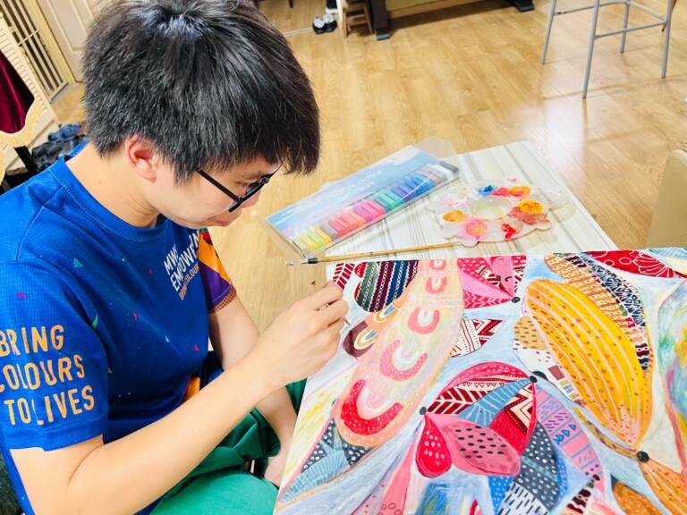 Young artist wearing glasses and a blue T-shirt seated at a table, carefully adding fine details to a vibrant mixed media painting. The artwork features butterfly-like shapes decorated with intricate geometric patterns, dots and stripes in pink, yellow, red and blue. Pastels, brushes and a paint palette are arranged on the table nearby.