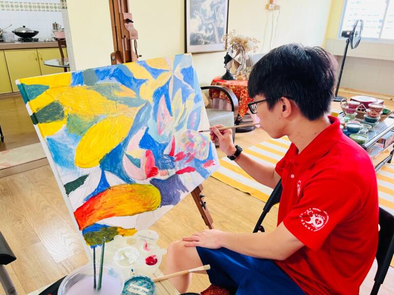 Young artist wearing glasses and a red polo shirt, seated at an easel and painting a vibrant abstract composition featuring bold yellow, blue, green, pink and orange shapes. Paintbrushes, palettes and art supplies are arranged nearby in a bright studio.