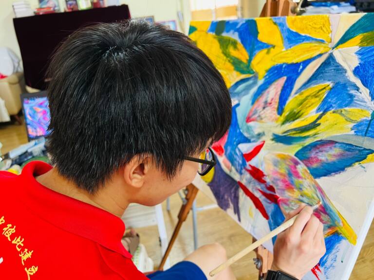 Close view of a young artist in glasses and a red polo shirt carefully painting an abstract canvas. The artwork features vibrant yellow, blue, green, pink and red shapes, with the artist adding intricate multicoloured brushstrokes to the lower right section.