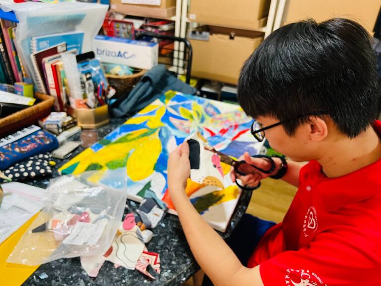 Young artist wearing glasses and a red polo shirt carefully cutting a piece of patterned fabric with scissors at a cluttered worktable. A vibrant abstract painting in yellow, blue, green and pink lies on the table, surrounded by fabric scraps, books and art supplies.