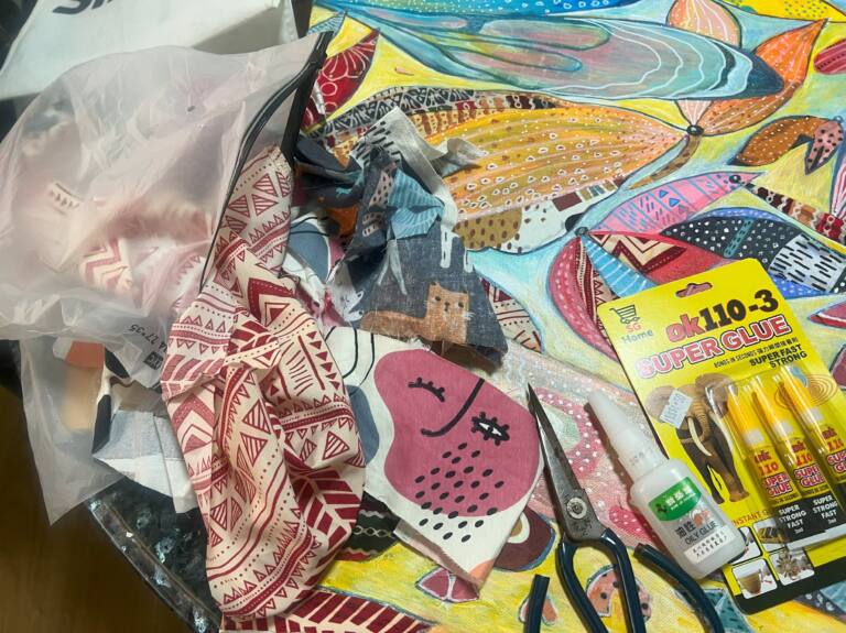 Overhead view of an artist’s workspace featuring colourful patterned fabric cuttings scattered across a vibrant abstract painting. Scissors, tubes of super glue and a bottle of fabric glue sit among the materials, showing the process of creating a mixed media artwork.
